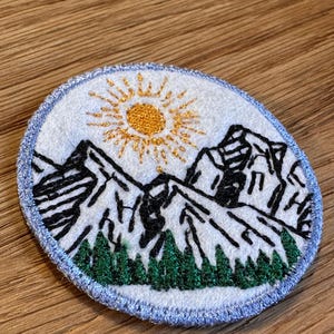 May include: A circular embroidered patch featuring a white background with a black stitched mountain range, a bright yellow sun with radiating rays, and a row of dark green pine trees at the base, all enclosed by a light blue stitched border.