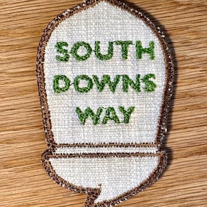 May include: A shield-shaped embroidered patch with a white fabric base and a brown stitched border. The patch features the green embroidered text SOUTH DOWNS WAY in a bold, sans-serif font. The design has a pointed top and a flat bottom edge.