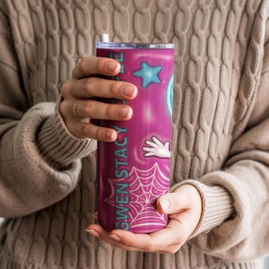 May include: A tall, cylindrical tumbler with a pink and purple gradient design. The tumbler features the name "GWEN STACY" in teal, along with cartoon graphics of a star, a hand, and a spiderweb. It has a silver lid and a white straw.