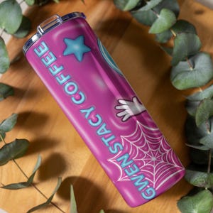 May include: A pink stainless steel tumbler with the words "GWEN STACY COFFEE" in light blue. The tumbler features a blue star, a white gloved hand, and a spiderweb design. The tumbler is on a wooden surface with green leaves.