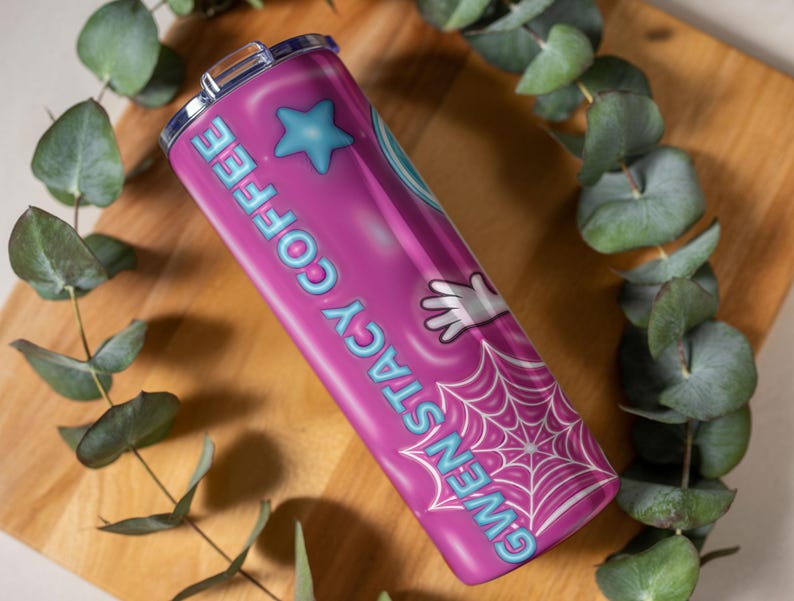 May include: A pink stainless steel tumbler with the words "GWEN STACY COFFEE" in light blue. The tumbler features a blue star, a white gloved hand, and a spiderweb design. The tumbler is on a wooden surface with green leaves.