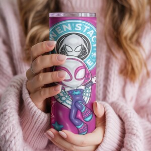 May include: A tall, cylindrical tumbler featuring a vibrant design of a superhero character. The tumbler is primarily pink and purple, with the character's image prominently displayed. The text "WEN STAC" is visible at the top. The tumbler is held by a person.