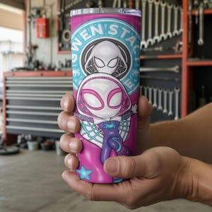 May include: A pink and teal tumbler featuring a Gwen Stacy Spider-Woman design. The tumbler has a white and pink illustration of the character, with the words "GWEN STACY" in a circular teal design. The tumbler is held in a person's hands.