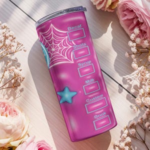 May include: A pink tumbler with a spiderweb design and a blue star. The tumbler has labels for drink customization, including "Decaf," "Shots," "Syrup," "Milk," "Custom," and "Drink." The tumbler is surrounded by pink roses and white flowers.