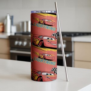 May include: A red tumbler featuring a repeating pattern of Lightning McQueen from the Cars movie. The design includes the number 95, a checkered flag, and the word "Lightning". A silver metal straw is next to the tumbler.