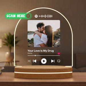 Peut inclure: Lampe LED avec base en bois et panneau acrylique en forme d'arche. Le panneau affiche une photo d'un couple, les détails d'une chanson et des commandes musicales. Le texte "SCAN HERE" est visible. Design décoratif.