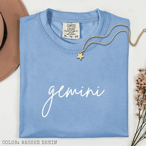 Pode incluir: Uma camiseta Comfort Colors azul claro com a palavra "Gemini" em escrita branca. Um colar dourado com um pingente de estrela repousa sobre a camiseta. Tamanho XL-TG-EG.