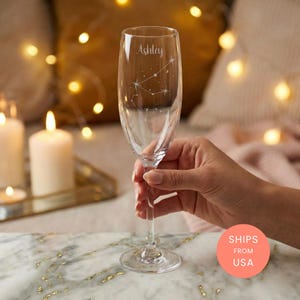 Op de afbeelding: Een heldere champagneflute met de naam "Ashley" en een sterrenbeeldontwerp geëtst op het glas. Het glas wordt door een hand vastgehouden. Op de achtergrond kaarsen en een marmeren oppervlak.