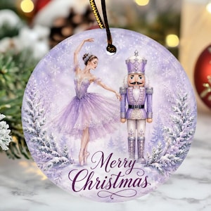 May include: A round ceramic Christmas ornament featuring a ballerina in a lavender tutu and a matching nutcracker standing in a snowy forest. The design includes snowflakes and the text Merry Christmas in a dark purple script font at the bottom.