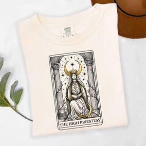 May include: A cream-coloured t-shirt with a tarot card design of The High Priestess. The illustration is in black and gold, showing a woman with a crescent moon crown, seated between pillars. The text "THE HIGH PRIESTESS" is at the bottom.