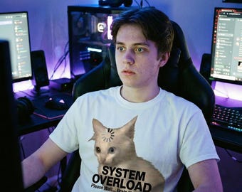 Könnte beinhalten: Ein weißes T-Shirt mit einem Katzen-Grafik und dem Text "SYSTEM OVERLOAD" und "Please Wait... Brain Not Found". Die Person sitzt vor einem Computer-Setup mit mehreren Bildschirmen und Peripheriegeräten.