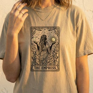 Pode incluir: Camiseta bege com um desenho preto de uma mulher segurando uma lua crescente e um sol, com o texto "THE EMPRESS" na parte inferior. O design é emoldurado por arabescos e elementos celestes, evocando uma carta de tarô.