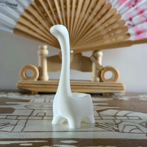 Peut inclure: Un porte-bague en forme de dinosaure en céramique blanche. Le dinosaure a un long cou et une petite zone ouverte sur son dos pour tenir les bagues. En arrière-plan, un éventail décoratif avec des motifs floraux roses et un support en bois.