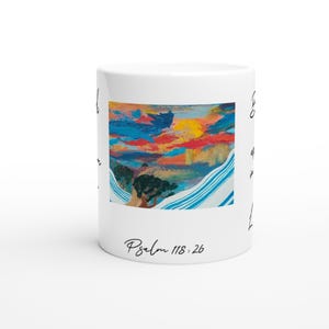 Peut inclure: Mug en céramique blanche orné d'une peinture de paysage colorée avec des tons de bleu, rouge, jaune et vert. Le mug porte l'inscription "Psalm 118:26". Le design comprend des accents rayés bleus et blancs.