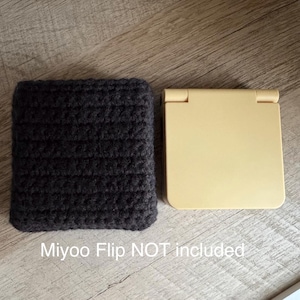 May include: A dark gray, crocheted sleeve next to a light yellow, square-shaped case. The sleeve is made of thick yarn and has a textured appearance. The case has a hinged top. The text "Miyoo Flip NOT included" is at the bottom.
