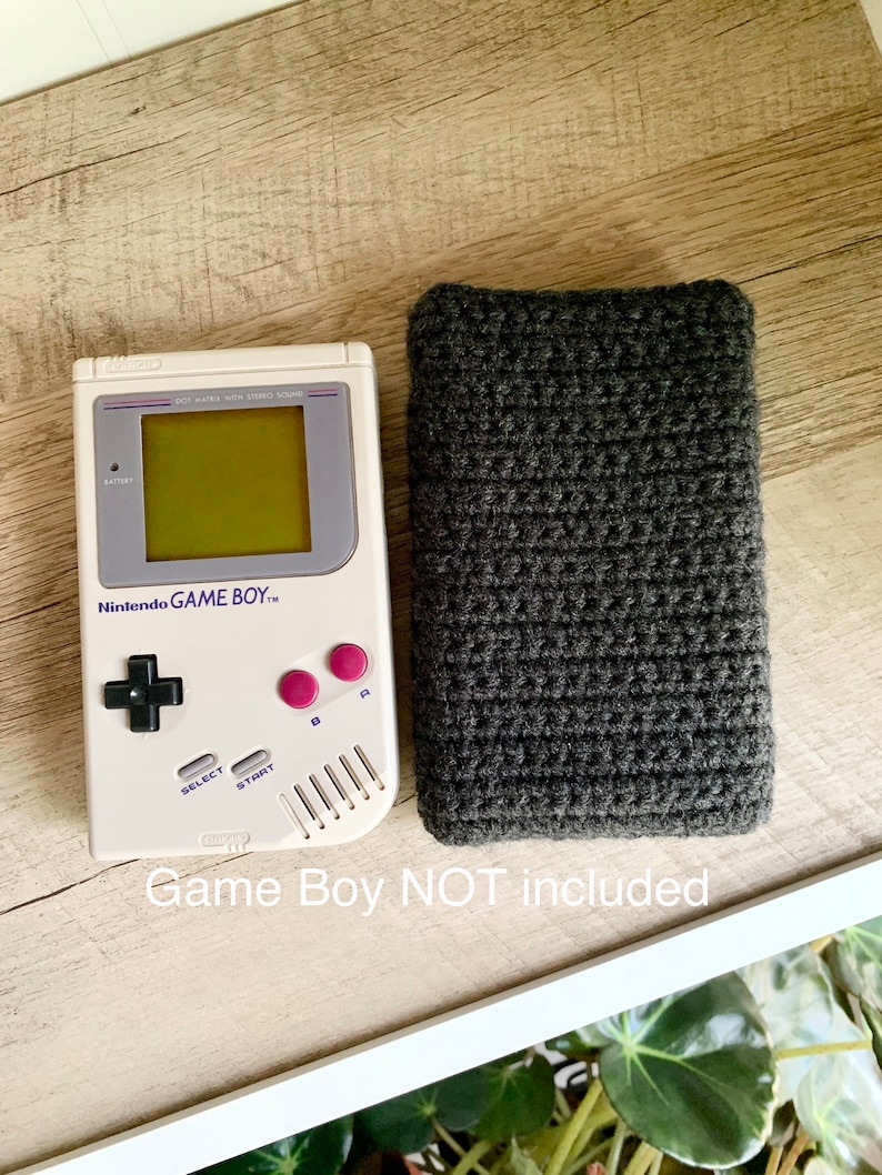 May include: A gray crocheted case for a Game Boy handheld video game console. The Game Boy is not included. The case is on a wooden surface.