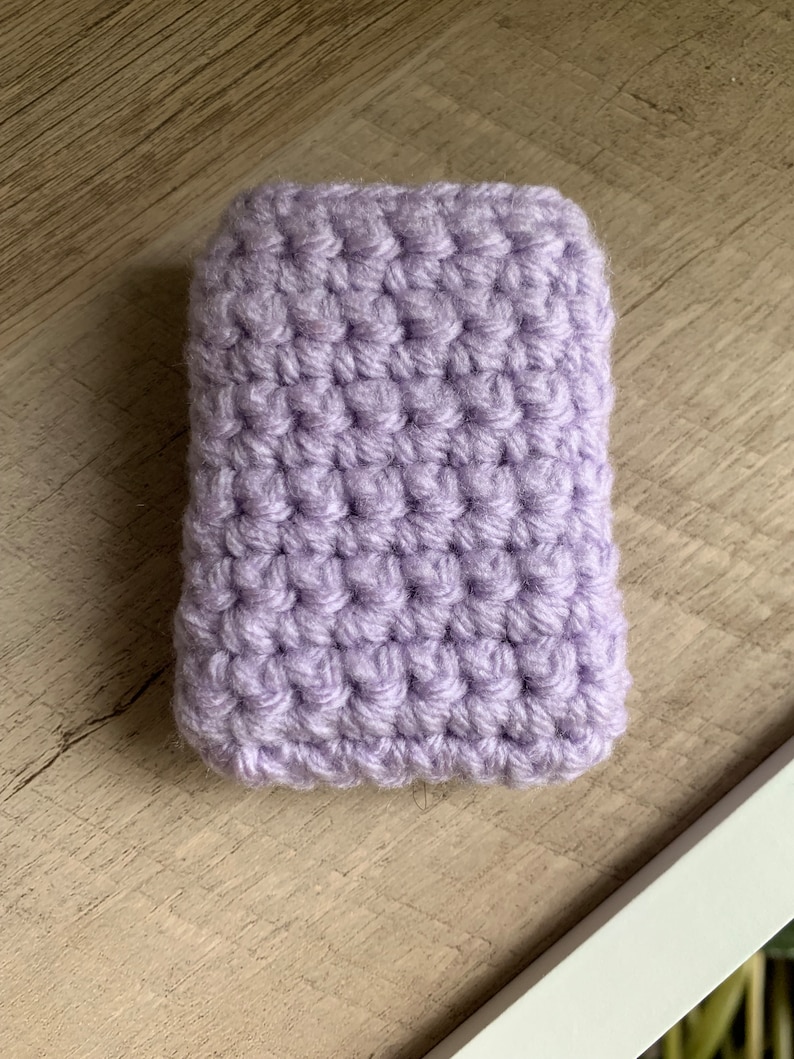 K&ouml;nnte beinhalten: Eine kleine, lilafarbene geh&auml;kelte Tasche. Die Tasche ist mit einem einfachen H&auml;kelmaschenstich gearbeitet und hat eine glatte, strukturierte Oberfl&auml;che.