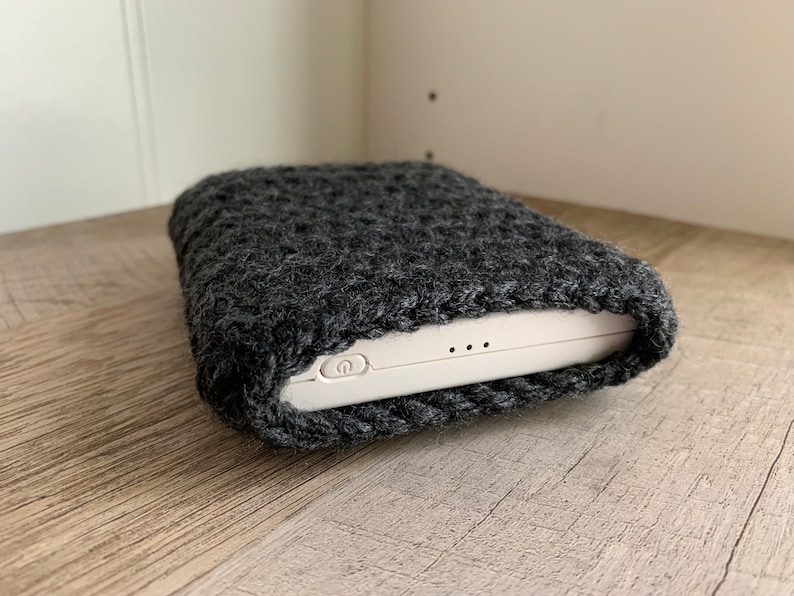K&ouml;nnte beinhalten: Eine graue Strickh&uuml;lle f&uuml;r ein wei&szlig;es elektronisches Ger&auml;t. Die H&uuml;lle besteht aus einem weichen, flauschigen Material und hat einen Rei&szlig;verschluss. Das Ger&auml;t hat einen Ein- / Ausschalter und zwei kleine L&ouml;cher an der Seite.