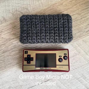 Puede incluir: Una funda de ganchillo gris y una consola Game Boy Micro roja y dorada. La funda es rectangular y parece hecha a mano. La Game Boy Micro tiene una pantalla, un mando direccional y botones A y B. El texto en la parte inferior dice "Game Boy Micro NOT included."