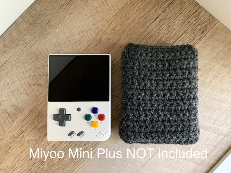 K&ouml;nnte beinhalten: Eine wei&szlig;e und graue Handheld-Videospielkonsole mit einem schwarzen Bildschirm und bunten Kn&ouml;pfen. Die Konsole liegt auf einer Holzoberfl&auml;che neben einer grauen geh&auml;kelten Tasche. Der Text "Miyoo Mini Plus NOT Included" steht unter dem Bild.