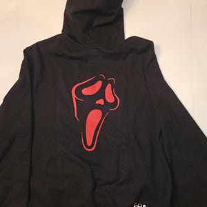 Könnte beinhalten: Schwarzer Kapuzenpullover mit roter Scream-Maske auf dem Rücken. Der Pullover hat eine Kapuze mit Kordelzug und lange Ärmel. Der Text "GoodieHoodie" ist unten rechts in Weiß aufgedruckt.