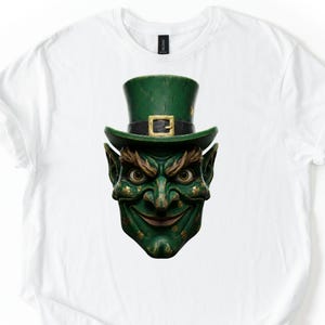 May include: White t-shirt featuring a detailed graphic of a green leprechaun face with a top hat. The leprechaun has a mischievous grin, pointed ears, and gold shamrocks. The hat has a gold buckle.
