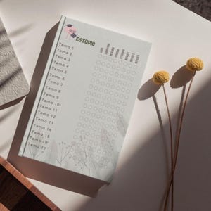 Puede incluir: Un cuaderno blanco con la palabra "ESTUDIO" y una ilustración de gorro de graduación de dibujos animados. El cuaderno tiene una lista de temas y casillas de verificación. Dos tallos de flores secas amarillas están en el fondo.