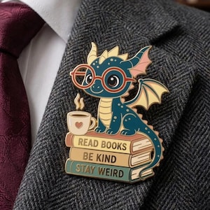 Pode incluir: Pin de esmalte de um dragão azul com óculos e uma chávena de café, pousado numa pilha de livros. Os livros dizem "READ BOOKS", "BE KIND" e "STAY WEIRD". O pin está preso a um blazer de tweed cinzento.