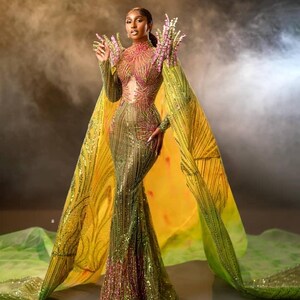 Puede incluir: Un glamuroso vestido largo con una silueta ajustada, adornado con lentejuelas en tonos verdes, dorados y rosas. El vestido presenta mangas largas y un cuello alto. Una capa fluida amarilla y verde se extiende desde los hombros, creando un efecto dramático.