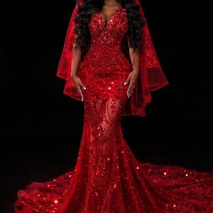 May include: A vibrant red, full-length gown with a low-cut neckline and a sheer, embellished skirt. The dress is adorned with sequins and has a long train. A matching red veil is draped over the shoulders, creating a dramatic effect.
