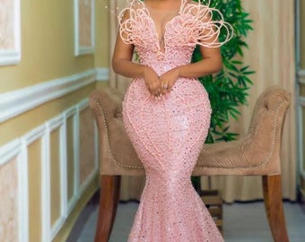 May include: A light pink, floor-length gown with a mermaid silhouette. The dress features a plunging neckline and is embellished with beads and sequins. The shoulders are adorned with elaborate, beaded accents. The gown has a long train.