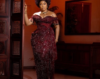 May include: A formal, off-the-shoulder burgundy gown with intricate beadwork and fringe detailing. The dress features a fitted bodice and a flowing skirt. A matching head wrap and jewelry complete the elegant ensemble.