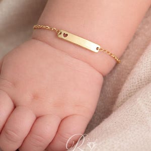 Könnte beinhalten: Ein goldfarbenes Babyarmband mit einem rechteckigen Namensschild und einem herzförmigen Ausschnitt. Das Armband ist an einer zarten Kette befestigt und wird am Handgelenk eines Babys gezeigt. Der Hintergrund ist in einer weichen, neutralen Farbe gehalten.