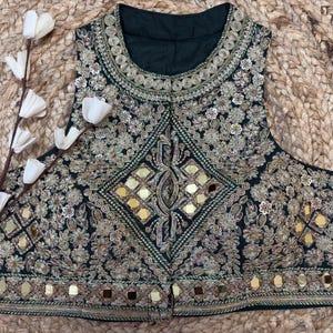 Puede incluir: Una blusa sin mangas de color verde oscuro con intrincados bordados dorados, patrones florales y detalles de espejos reflectantes en la parte delantera. La prenda tiene un cuello redondo alto y una banda horizontal de espejos cuadrados en el dobladillo.