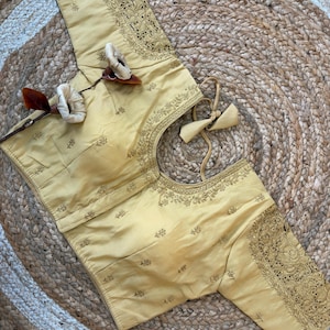 May include: A pale gold blouse with detailed embroidery on the sleeves and neckline. The blouse has a round neck and long sleeves. The embroidery is a darker gold, contrasting with the lighter fabric. The blouse is displayed on a woven surface, likely a rug or mat.