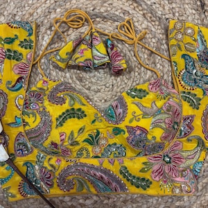 May include: A vibrant yellow embroidered top with floral and paisley designs in pink, green, and blue. The top has a V-neck, short sleeves, and decorative tassels. The fabric has a subtle sheen, and the design is detailed with small embellishments.