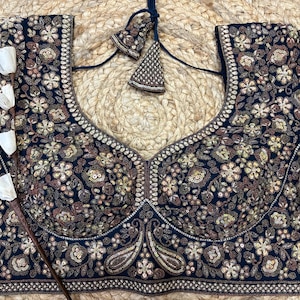 Può includere: Una camicetta blu navy a maniche corte con ricami floreali intricati nei toni del marrone e dell'oro. La camicetta presenta una profonda scollatura a V ed è impreziosita da paillettes. Un paio di nappe decorative abbinate pendono dallo scollo.