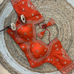 Puede incluir: Una blusa naranja con bordados y adornos plateados. La blusa presenta un escote corazón y se combina con mangas a juego. El diseño incluye detalles florales y en forma de estrella, creando un look elegante. La prenda se exhibe sobre una alfombra tejida.