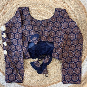 May include: A navy blue blouse with a repeating floral pattern in shades of brown and white. The blouse has long sleeves and a scoop neckline. The back of the blouse is open, revealing a dark blue underlay with tie closures. The blouse is displayed on a woven mat.