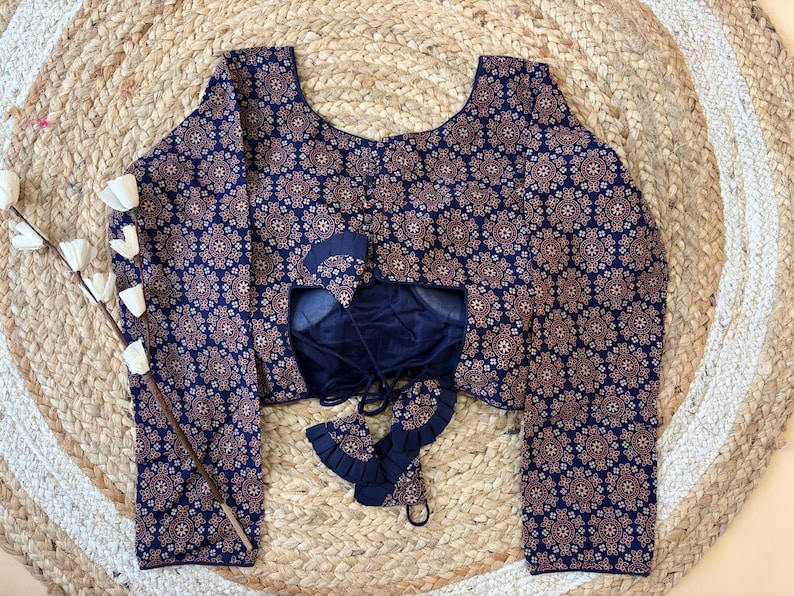 May include: A navy blue blouse with a repeating floral pattern in shades of brown and white. The blouse has long sleeves and a scoop neckline. The back of the blouse is open, revealing a dark blue underlay with tie closures. The blouse is displayed on a woven mat.