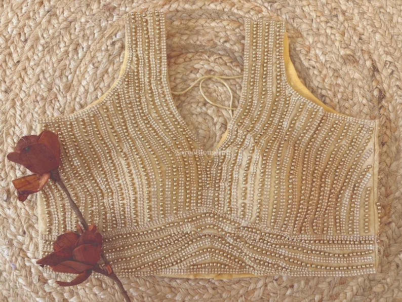 May include: A sleeveless, gold-colored beaded top with a deep V-neck. The intricate beadwork creates vertical lines across the bodice. A pair of dark red artificial flowers are placed on the left side of the top.