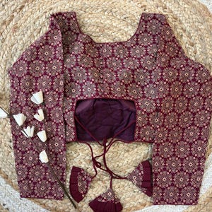 May include: A maroon blouse with a repeating white floral pattern. The long-sleeved top has a scoop neckline and a keyhole back. The blouse has a drawstring closure and decorative tassels. The blouse is displayed on a woven, circular rug.
