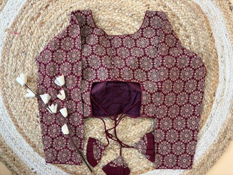 May include: A maroon blouse with a repeating white floral pattern. The long-sleeved top has a scoop neckline and a keyhole back. The blouse has a drawstring closure and decorative tassels. The blouse is displayed on a woven, circular rug.
