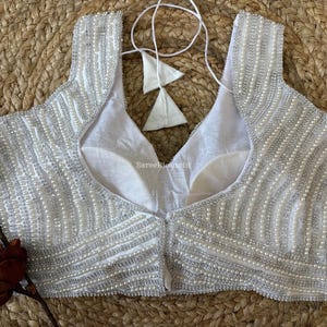 May include: A white, sleeveless top with intricate beadwork. The garment features a scoop neckline and is adorned with rows of small, silver-colored beads. The back of the top is plain white, and it has a tie closure. The top is likely a blouse or a formal wear item.