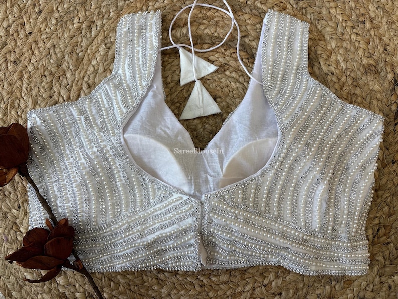May include: A white, sleeveless top with intricate beadwork. The garment features a scoop neckline and is adorned with rows of small, silver-colored beads. The back of the top is plain white, and it has a tie closure. The top is likely a blouse or a formal wear item.
