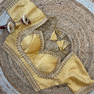May include: A gold-colored blouse with intricate embroidery and sequin detailing. The blouse features a scoop neckline and short sleeves. The garment is displayed on a woven, circular mat.
