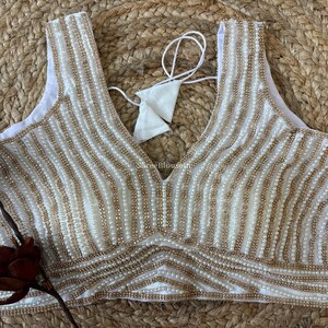 May include: White sleeveless top with a deep V-neckline, embellished with vertical rows of small, gold-colored beads. The beaded design covers the entire front of the top. A white tie is visible at the back.