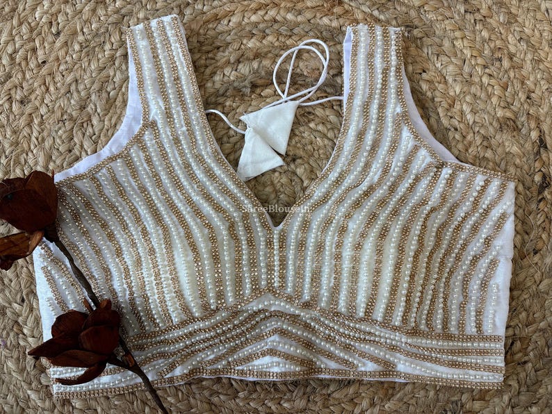May include: White sleeveless top with a deep V-neckline, embellished with vertical rows of small, gold-colored beads. The beaded design covers the entire front of the top. A white tie is visible at the back.