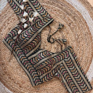 May include: Black embroidered top with gold, orange, and pink leaf pattern. The top is embellished with gold sequins and has a black tie closure. The garment is displayed on a woven jute rug.