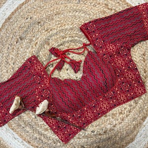 May include: A red and black patterned top with matching sleeves and a tie-up back. The top is made of a fabric with a geometric design and is displayed on a woven, circular mat.